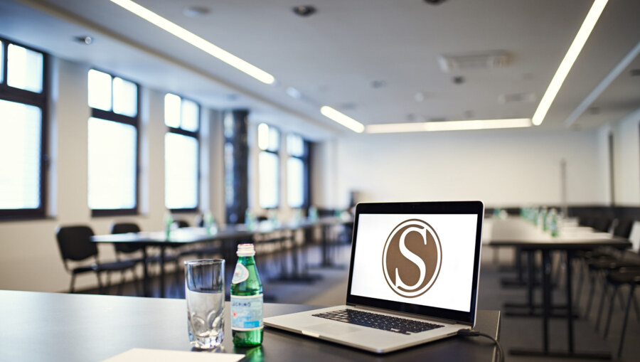 Open laptop with 'S' logo, water bottle and empty glass on desk, long meeting tables visible behind.