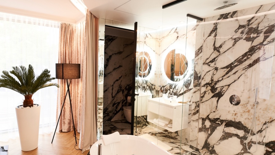 Bright marble bathroom featuring a white bathtub, glass-enclosed shower, glowing circular mirrors, and soft curtains.
