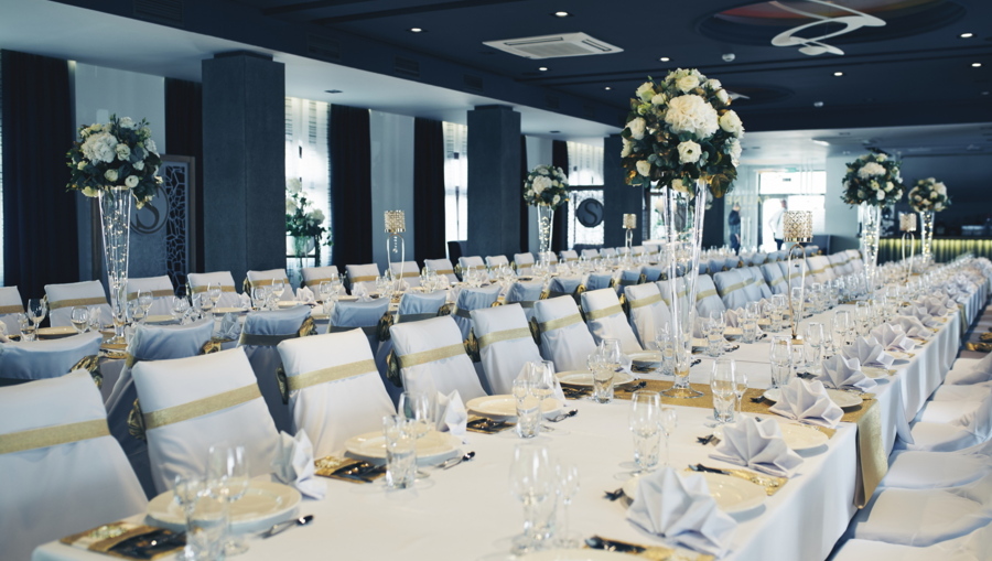 Long banquet tables dressed in white and gold, repeated tall floral centerpieces, elegant formal setting.