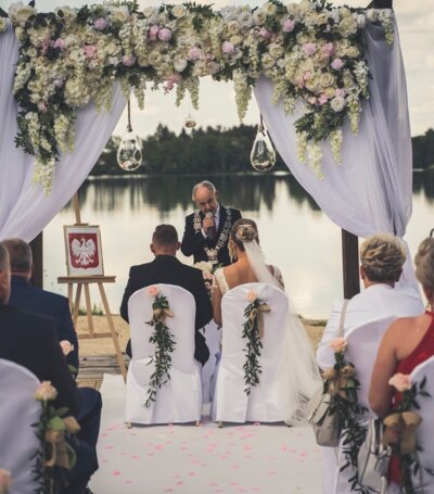 Ceremonia ślubna na tle jeziora z kwiatową dekoracją, białymi pokrowcami na krzesłach i uczestnikami w eleganckich strojach