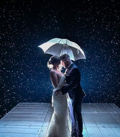Para nowożeńców w eleganckim stroju podświetlona parasolką na drewnianym tarasie podczas nocnego deszczu