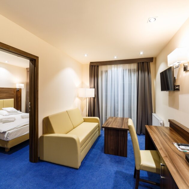 Przytulny apartament z jasnym salonem, beżową sofą, drewnianym biurkiem, szafką z czajnikiem i oddzielną sypialnią z dużym łóżkiem.