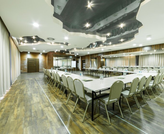 Nowoczesna sala konferencyjna z drewnianą podłogą, okrągło-kwadratowymi stołami, krzesłami i oświetleniem sufitowym odbijającym się w lustrze