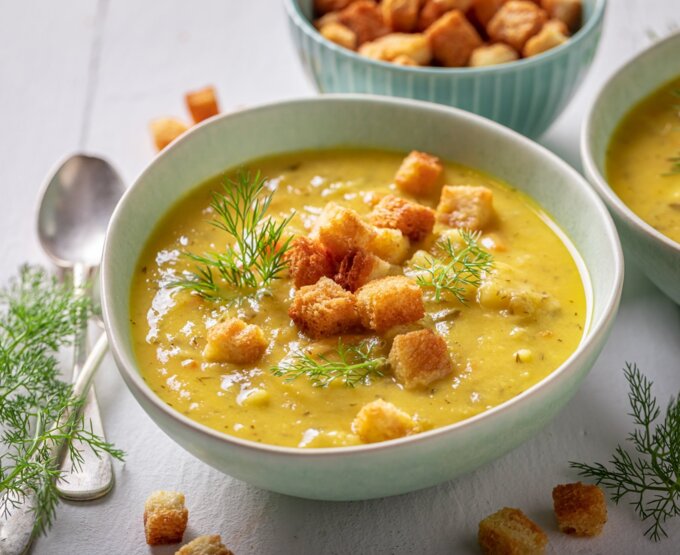 Zupa krem z warzyw w misce, podana z chrupiącymi grzankami i świeżym koperkiem, na białym stole z łyżkami obok