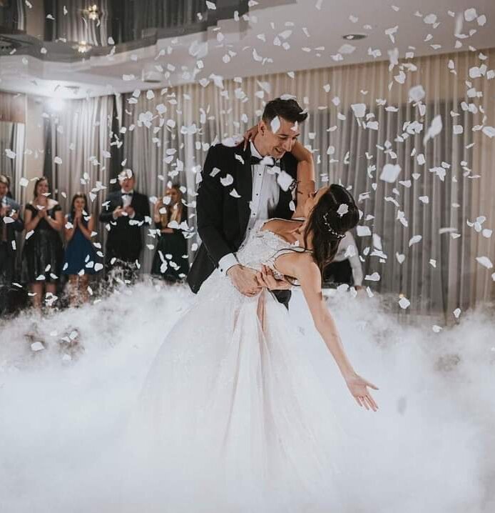 Para młoda tańczy pierwszy taniec na sali weselnej z efektem mgły i wirującymi płatkami kwiatów, goście obserwują