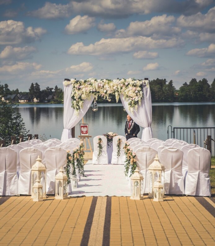 Plenerowa aranżacja ślubna nad jeziorem z białymi krzesłami, kwiatowym baldachimem i ozdobnymi latarniami przy słonecznej pogodzie