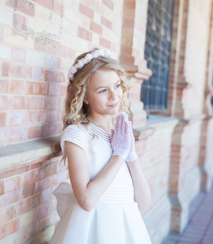 Młoda dziewczynka w białej sukni komunijnej i koronie kwiatowej stoi przy ścianie z cegieł w kolonadowym przejściu.