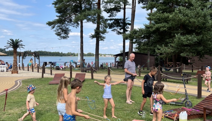 Dzieci bawią się na zielonej trawie nad jeziorem, w tle leżaki, palmy i plaża z plażowiczami w słoneczny dzień