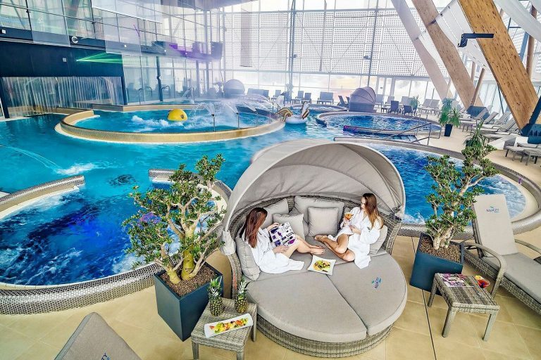 Two women in white robes relax on a covered lounge chair by the indoor pool with whirlpools and fountains in a luxury spa.