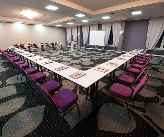 Sala konferencyjna w układzie U z fioletowymi krzesłami, białymi stołami, ekranem projekcyjnym i flipchartem, wzorzystym dywanem i miękkim oświetleniem