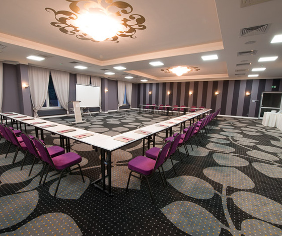 Sala konferencyjna w układzie U z fioletowymi krzesłami, pulpitem, ekranem projektora, wzorzystym dywanem i stolikiem z napojami