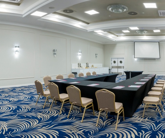 Sala konferencyjna w układzie U z ekranem projekcyjnym, czarne obrusy, beżowe krzesła, bufet kawowy i niebieski wzorzysty dywan