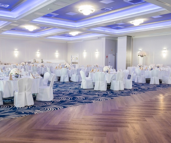 Elegancka sala bankietowa przygotowana na wesele: okrągłe stoły w bieli, kwiatowe kompozycje, niebieskie podświetlenie i parkiet