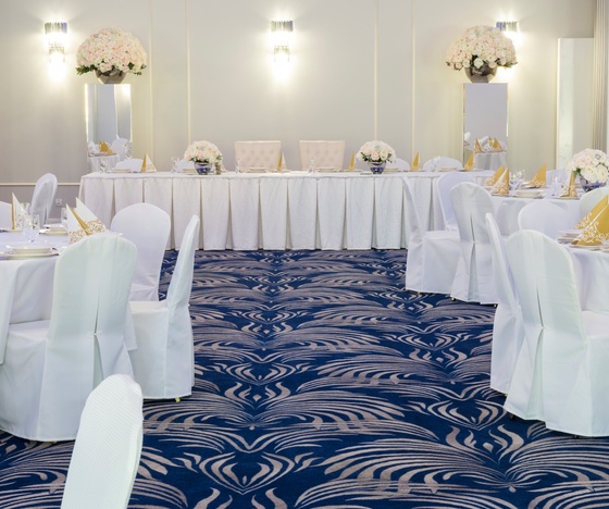 Elegancka sala bankietowa z okrągłymi stołami, białymi obrusami, złotymi serwetkami, bukietami róż i głównym stołem pod miękkim światłem.