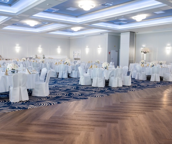 Elegancka sala weselna z okrągłymi stołami w białych obrusach, kwiatowymi dekoracjami, złożonymi serwetkami i drewnianym parkietem.