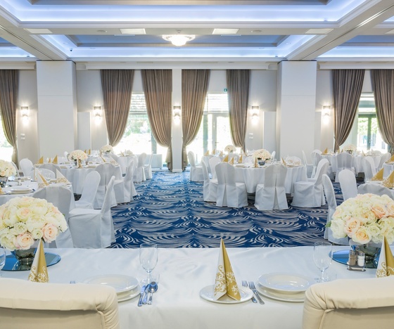 Elegancka sala bankietowa w hotelu przygotowana na wesele: okrągłe stoliki w białych pokrowcach, złote serwetki, bukiety róż, wysokie okna z zasłonami