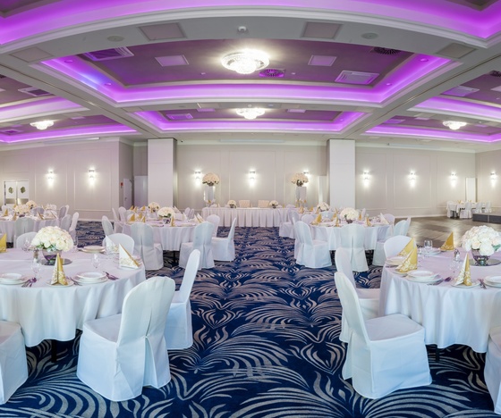 Elegancka sala bankietowa na wesele, okrągłe stoły w białych obrusach, złote serwetki, kwiatowe dekoracje, fioletowe oświetlenie sufitowe