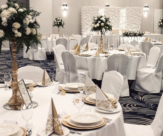 Elegancka sala bankietowa na wesele z okrągłymi stołami, białymi pokrowcami, złotymi nakryciami i wysokimi bukietami