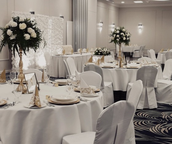 Elegancka sala bankietowa na przyjęcie ślubne z okrągłymi stołami w białych obrusach, złotymi akcentami i wysokimi bukietami białych róż