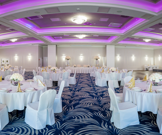 Elegancka sala bankietowa z okrągłymi stołami w białych pokrowcach, kwiatowymi dekoracjami, złotymi serwetkami, fioletowym oświetleniem sufitowym i niebieskim dywanem
