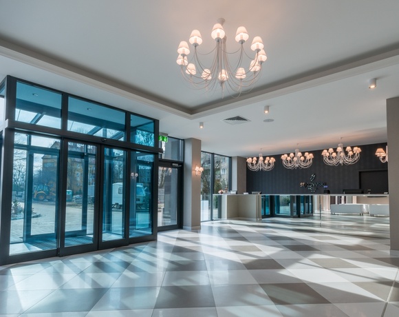 Jasne lobby z przeszklonym wejściem, szachownicową posadzką, eleganckimi żyrandolami i nowoczesną recepcją