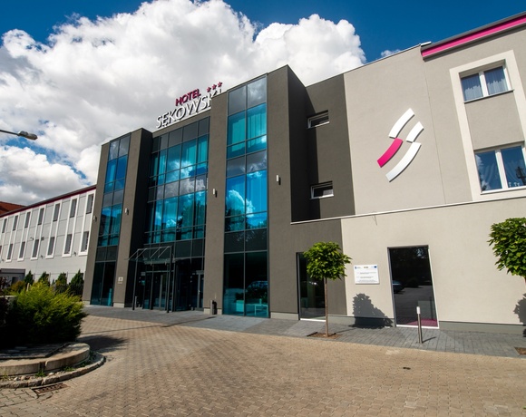 Nowoczesny trzykondygnacyjny hotel z przeszklonym wejściem, brukowanym podjazdem, małymi drzewkami i zadaszonym miejscem wypoczynkowym.