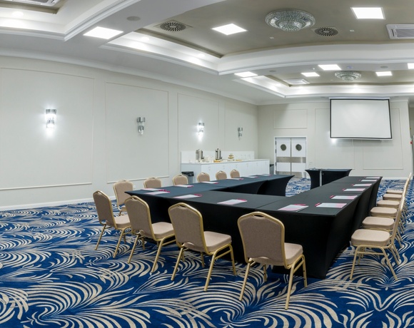 Sala konferencyjna w układzie U z ekranem projekcyjnym, czarne obrusy, beżowe krzesła, bufet kawowy i niebieski wzorzysty dywan