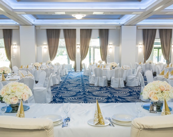 Elegancka sala bankietowa w hotelu przygotowana na wesele: okrągłe stoliki w białych pokrowcach, złote serwetki, bukiety róż, wysokie okna z zasłonami
