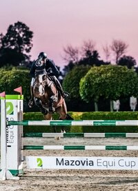 Wieczorny konkurs skoków: jeździec i koń przeskakują przeszkodę z napisem Mazovia Equi Cup na oświetlonej arenie.