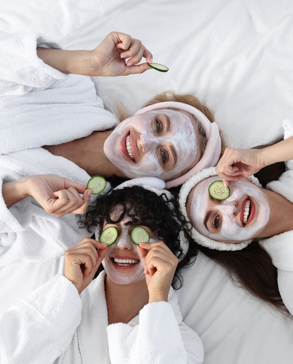 Trzy uśmiechnięte kobiety w białych szlafrokach z maseczkami i plasterkami ogórka, relaks w hotelowym spa