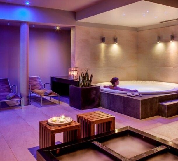 Relaksujące spa wewnętrzne z podświetlanym jacuzzi, kobietą w kąpieli, leżakami, drewnianymi stolikami, świecami i minimalistycznym wnętrzem.
