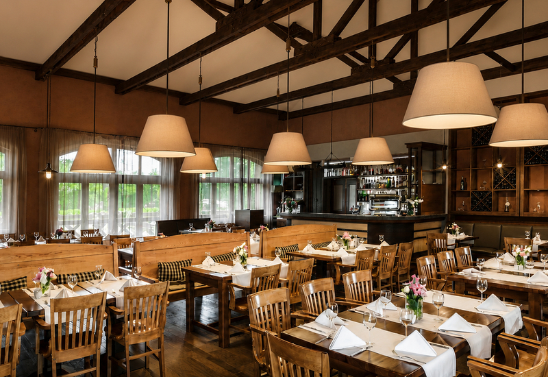 Przytulna restauracja z drewnianymi stolikami, nakrytymi białymi serwetkami, barem z regałem na wino i dużymi lampami.