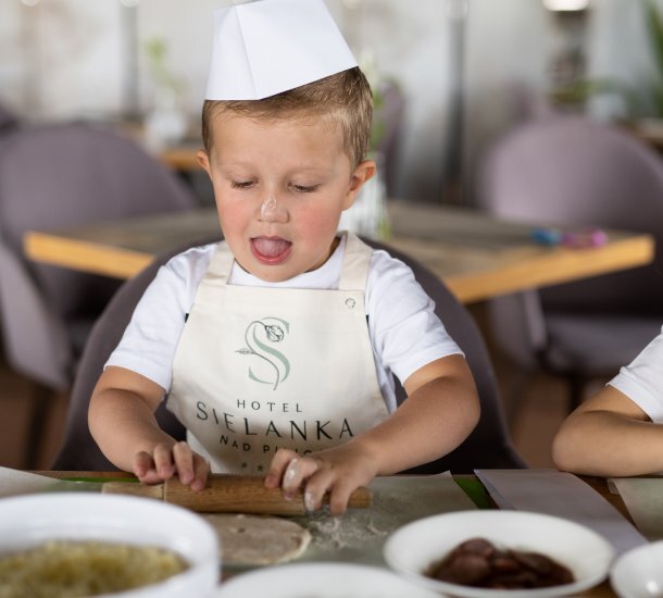 Dziecko w kucharskim czepku i fartuszku hotelu Sielanka wałkuje ciasto podczas zajęć kulinarnych w restauracji