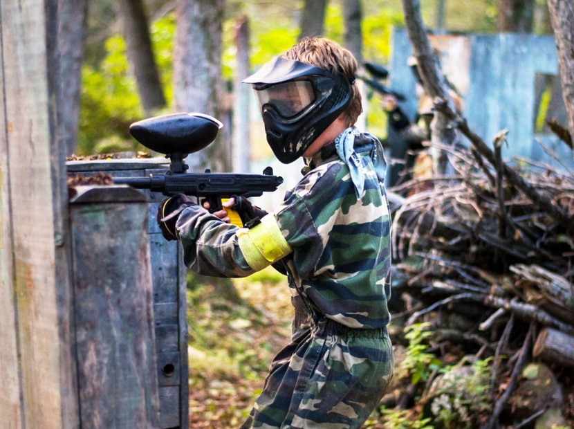 Gracz paintballowy w kamuflażu i masce, osłania się przy drewnianej barykadzie na leśnym polu paintballowym