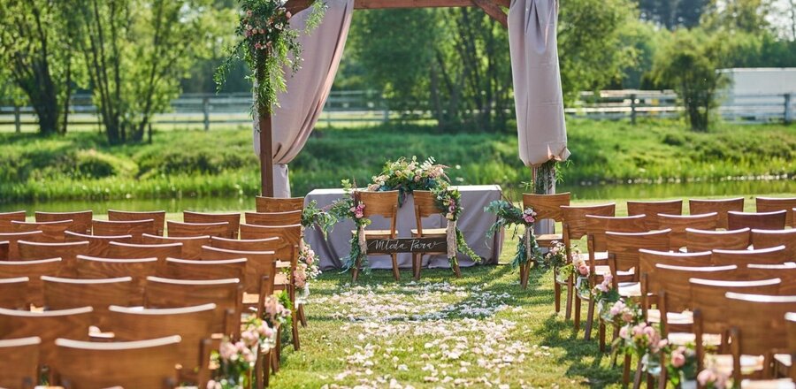 Plenerowa ceremonia ślubna nad stawem — rzędy drewnianych krzeseł, kwiatowa brama, alejka z płatków i stół pary młodej