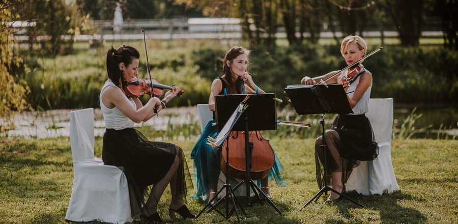 Trio smyczkowe gra na zewnątrz w ogrodzie hotelowym przy stawie, białe pokrowce na krzesłach, elegancka, romantyczna oprawa ślubna.