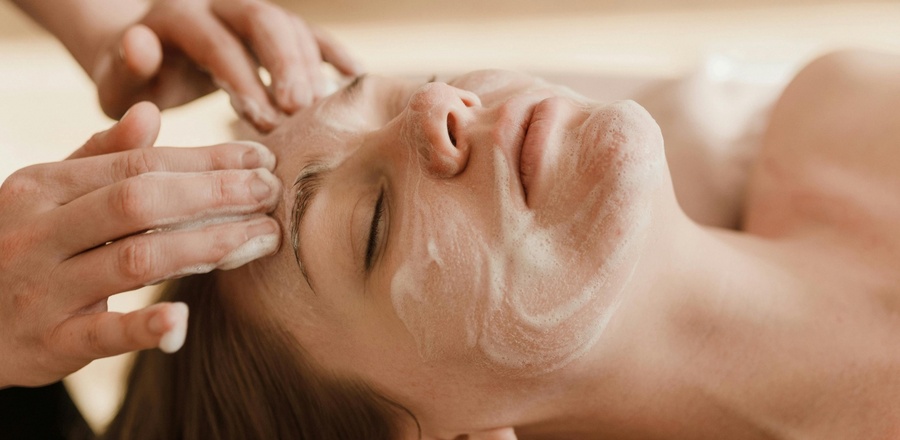 Relaksujący zabieg spa: kobieta z zamkniętymi oczami podczas oczyszczającego masażu twarzy z pianką.