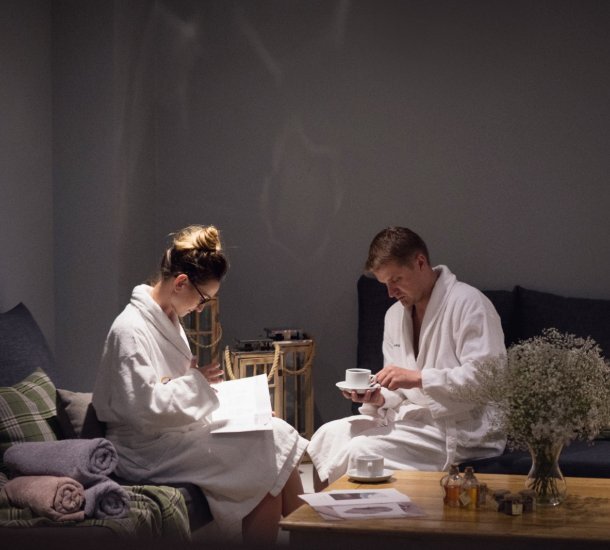 Dwoje gości w białych szlafrokach w przytulnym salonie spa, miękkie fotele, zrolowane ręczniki, filiżanki herbaty i wazon z gipsówką.