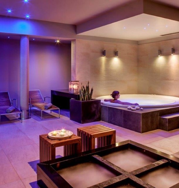 Relaksujące spa wewnętrzne z podświetlanym jacuzzi, kobietą w kąpieli, leżakami, drewnianymi stolikami, świecami i minimalistycznym wnętrzem.