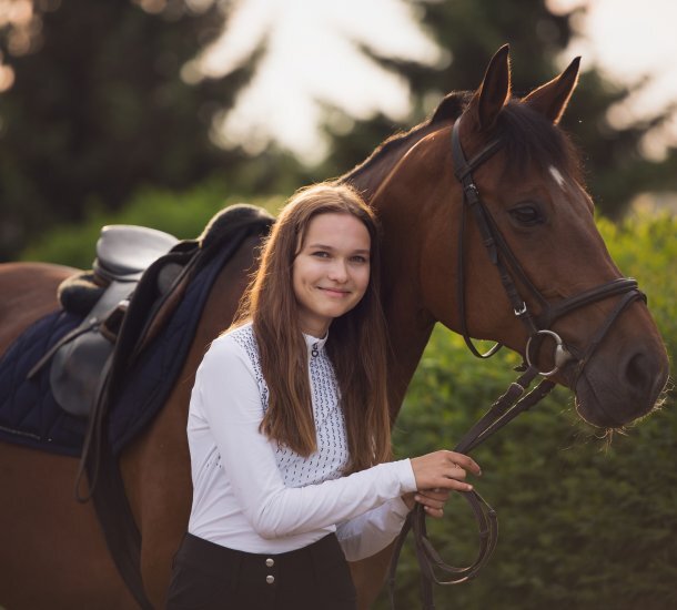 Uśmiechnięta młoda kobieta stojąca przy siodłanym koniu na tle zieleni, gotowa do jazdy