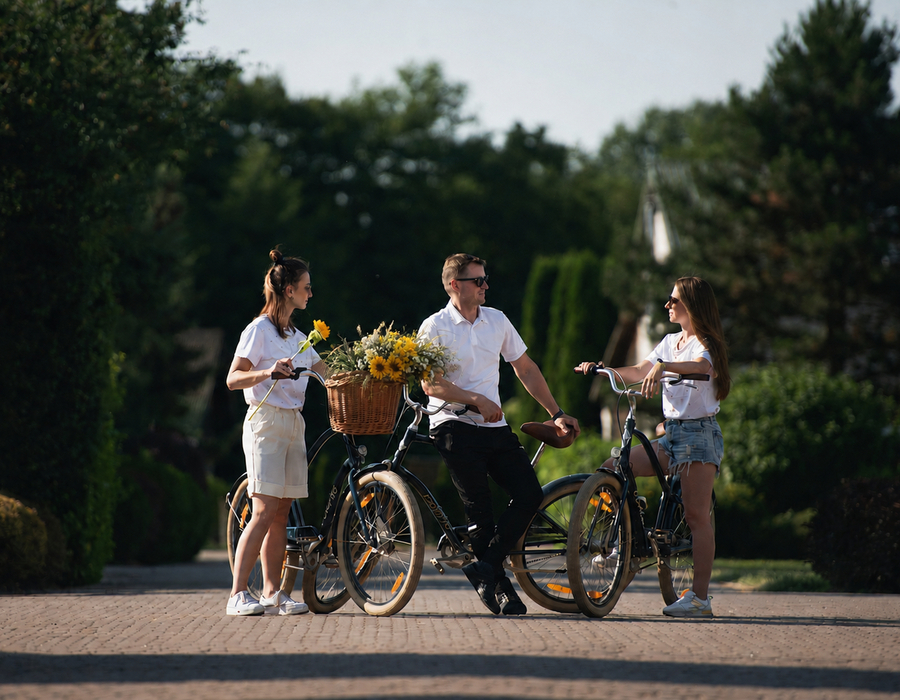 Trójka gości na rowerach na hotelowej alei — kosz z żółtymi kwiatami, letni, relaksujący klimat w zielonym ogrodzie