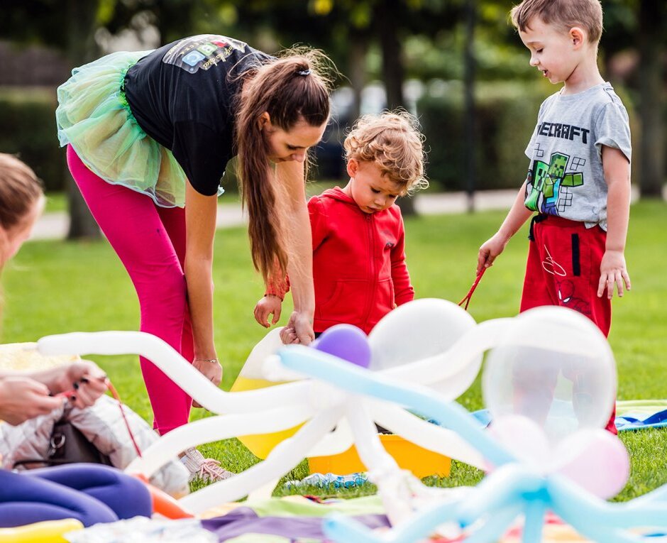 Animatorka i dzieci tworzą figurki z balonów na trawniku, kolorowe koce i stroje tworzą radosną, rodzinną atmosferę