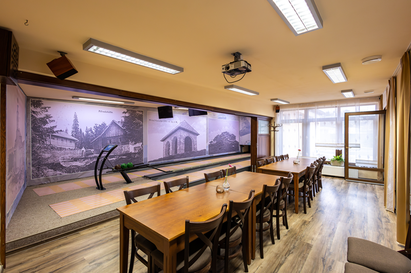 Hotelová kuželna s dvěma dřevěnými stoly, sezením pro skupiny, projektorem, historickými nástěnnými snímky a velkými okny