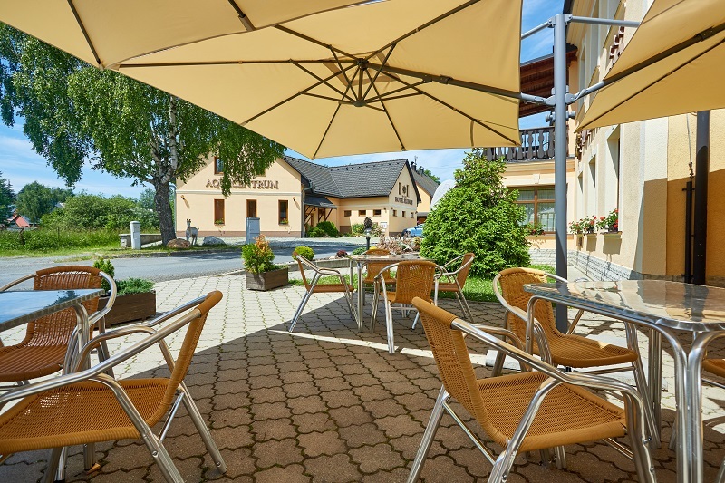 Zastíněná hotelová terasa s ratanovými židlemi a kovovými stolky pod velkými slunečníky, posezení venku s výhledem do zahrady a sousedního penzionu.