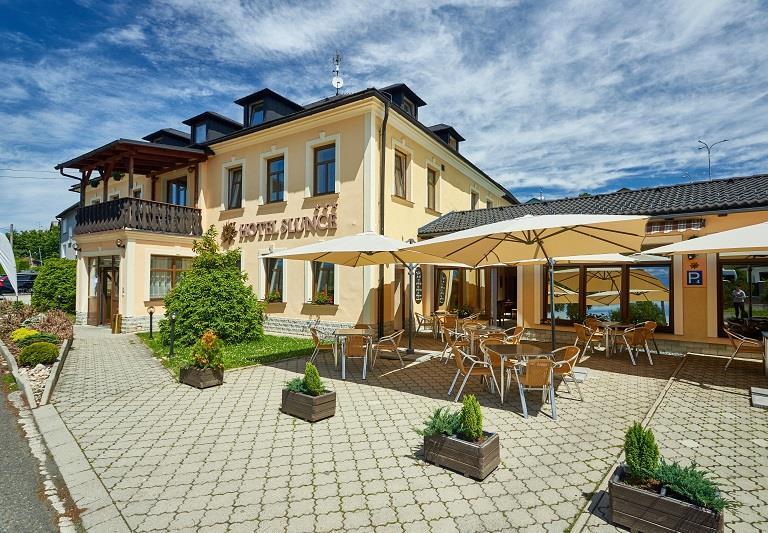 Dvoupodlažní žlutý hotel s dřevěným balkonem a slunnou dlážděnou terasou s stolky, slunečníky a květinami v květináčích.