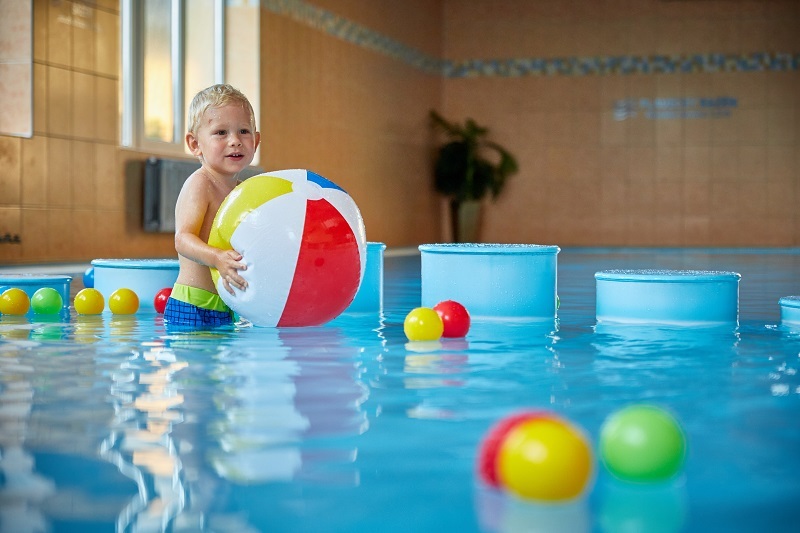 Malé dítě si hraje s velkým barevným plážovým míčem v mělkém krytém hotelovém bazénu se plovoucími hračkami.
