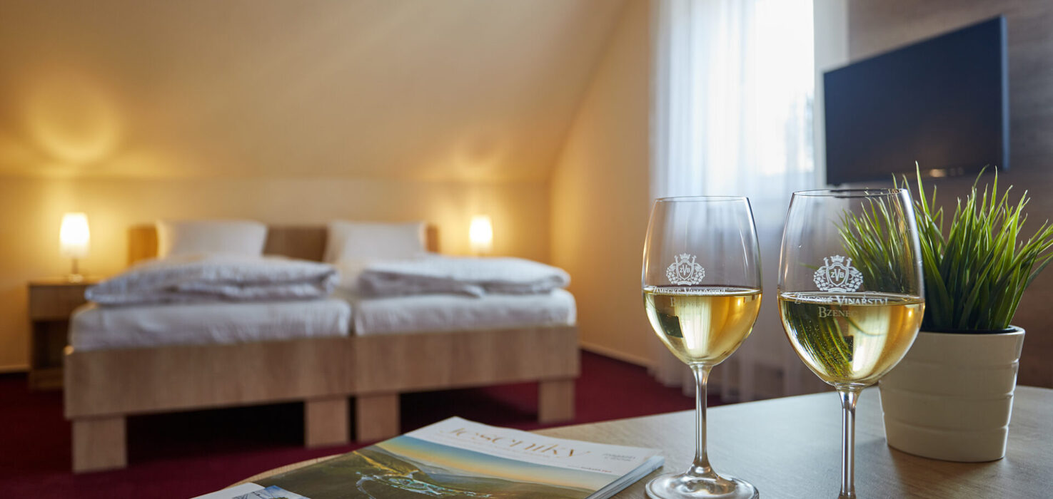 Útulný dvoulůžkový pokoj v Hotelu Slunce v Rýmařově v Jeseníkách s tlumeným světlem a příjemnou atmosférou. Na stolku stojí dvě sklenice bílého vína, časopisy a pokojová rostlina. Moderní a pohodlné ubytování v Jeseníkách ideální pro romantický nebo relaxační pobyt.