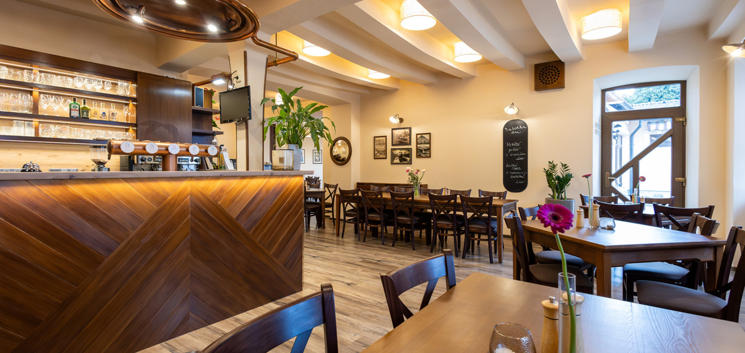 Příjemná hotelová jídelna s dřevěným barem, měděným krytem nad barem, dlouhými společnými stoly, odkrytými trámy a růžovým květem na stole.