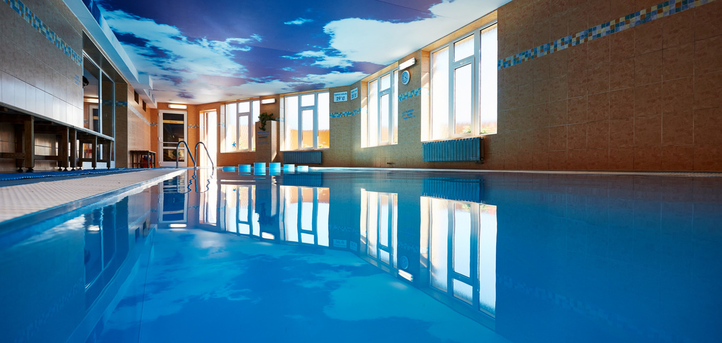 Vnitřní hotelový bazén s nebeskou malbou na stropě, velkými okny, dlaždicovým okrajem a zrcadlící klidnou hladinou.