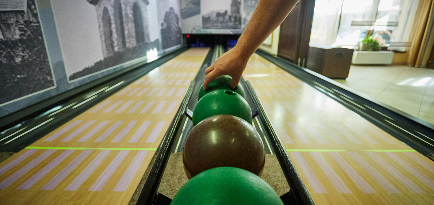 Privátní hotelová bowlingová dráha s dřevěnými dráhami a řadou zelených a hnědých koulí, host sahá po kouli